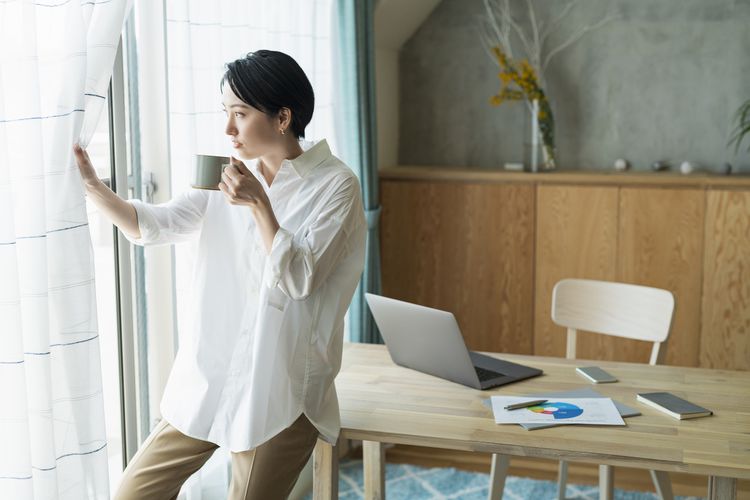A person with short black hair leans against a table, drinking from a mug while looking out a window