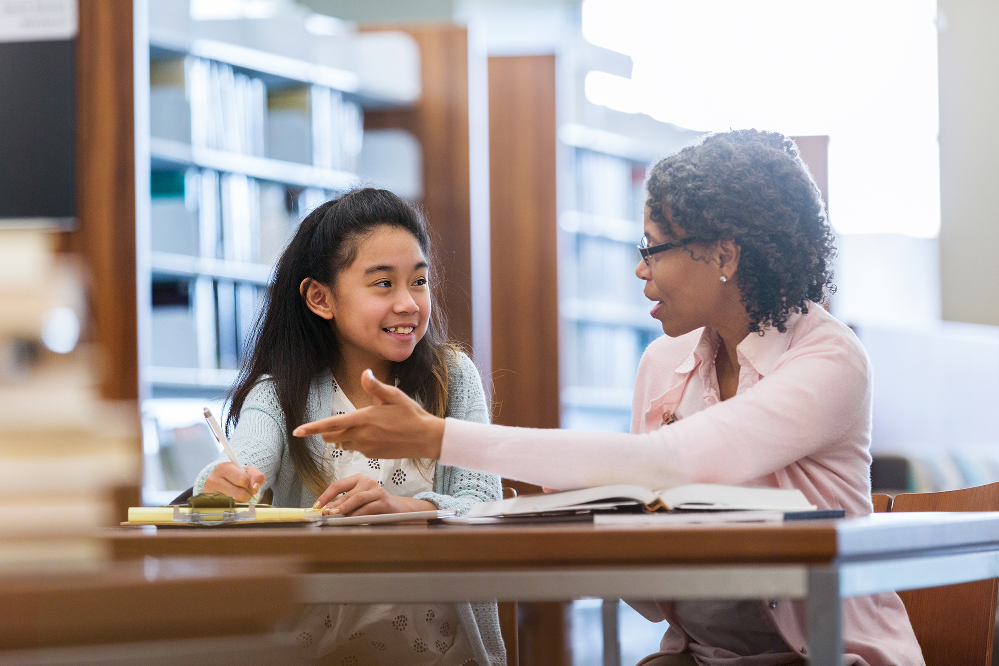 A smiling elementary age girl sits with her mature female mentor in a school library. She enjoys a conversation.